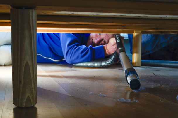 A man vacuums dust under a bed in an apartment - Photo, Image