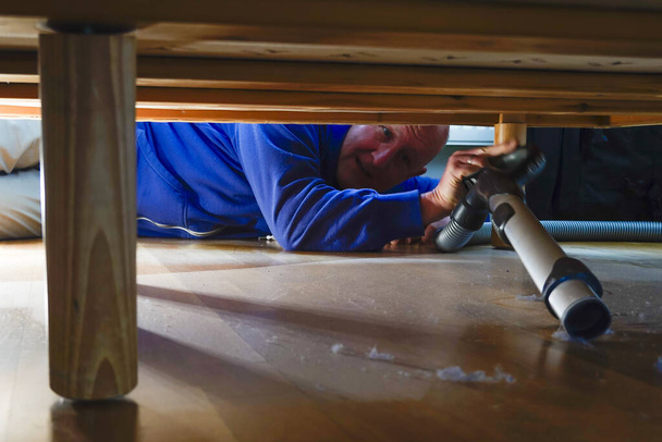 Um homem aspira poeira sob uma cama em um apartamento
 - Foto, Imagem