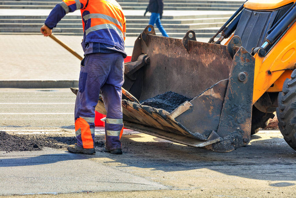 Ein Straßenmeisterei-Techniker schüttet während der Straßenbauarbeiten frischen Asphalt aus einem Eimer einer Planierraupe. - Foto, Bild