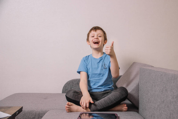 Щасливий веселий хлопчик грає на планшеті, сидячи на дивані під час ізоляції
 - Фото, зображення