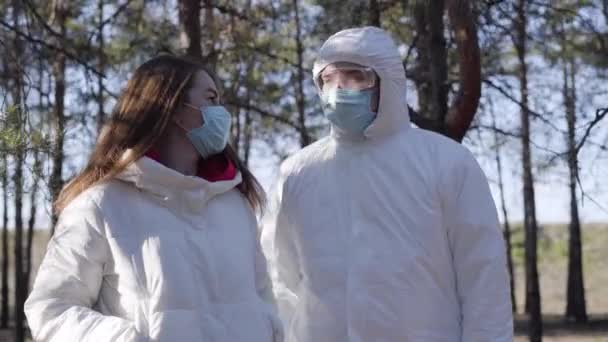 Young Caucasian man and woman in face masks talking to each other and turning to camera. Couple wearing safety suit outdoors on coronavirus quarantine. Covid-19 global pandemic, lifestyle. - Filmati, video