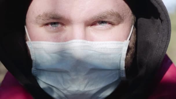 Extreme close-up face of young Caucasian man in face mask on sunny day outdoors. Portrait of confident handsome virologist looking at camera. Covid-19, coronavirus, quarantine, pandemic, lifestyle. - Video