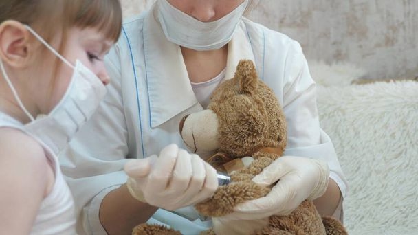 Χαριτωμένο κορίτσι κάνει μια ένεση για να παίξει αρκούδα. Το μωρό παίζει με ιατρική μάσκα. Το παιδί παίζει στο νοσοκομείο. παιχνίδι προσποιείται ότι είναι γιατρός, νοσοκόμα, κτηνίατρος, θεραπεύει έναν ασθενή με ένα εμβόλιο. Κίνδυνος κορωναϊού - Φωτογραφία, εικόνα