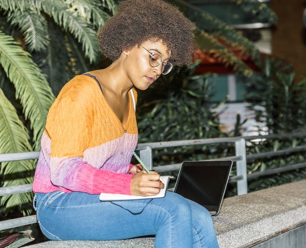 Mladá žena s brýlemi a afro vlasy psaní do notebooku sedí uvnitř stanice - Fotografie, Obrázek
