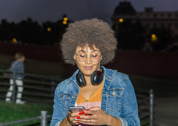 Koyu tenli, saçı kıvırcık, boynunda kulaklıkla akıllı telefonuna bakan genç bir kadının portresi. - Fotoğraf, Görsel