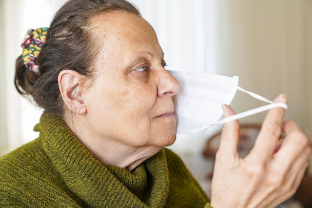 Портрет старої жінки в хірургічній масці для захисту від коронавірусу. Бабуся сидить у вітальні і дивиться вгору
  - Фото, зображення