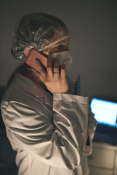 Doctor making a phone call. Hospital staff working at night duty. Woman wearing uniform, cap and face mask to prevent virus infection - Foto, Bild