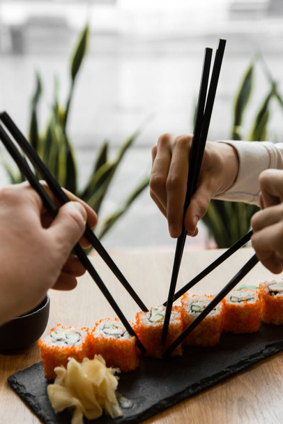 Οι άνθρωποι χρησιμοποιούν ξυλάκια για να φάνε σούσι. Πολλά χέρια με ξυλάκια τρώνε ρολό σούσι. - Φωτογραφία, εικόνα