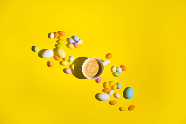 A lot of multi-colored Easter eggs and a mug of coffee with froth on a yellow background. Happy Easter card with copy space for text. Minimal style. In a hard light. selective focus. - Foto, immagini