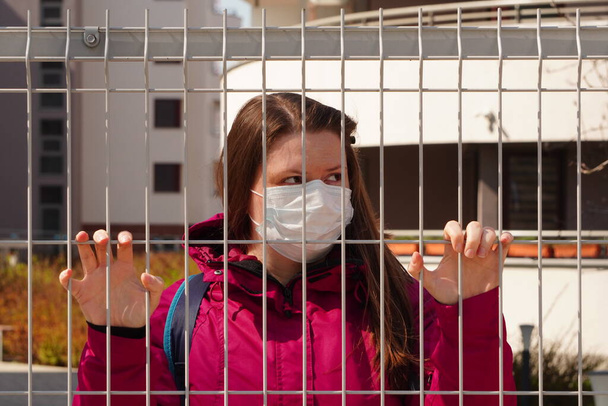 Tıbbi maske takmış açık tenli ve koyu renk saçlı genç bir kadın ya da kız çitin arkasında duruyor. Pandemik koronavirüs sırasında karantinadan kaçmak. İnsan sağlığı güvenliği. Giriş ve çıkış yasağı - Fotoğraf, Görsel
