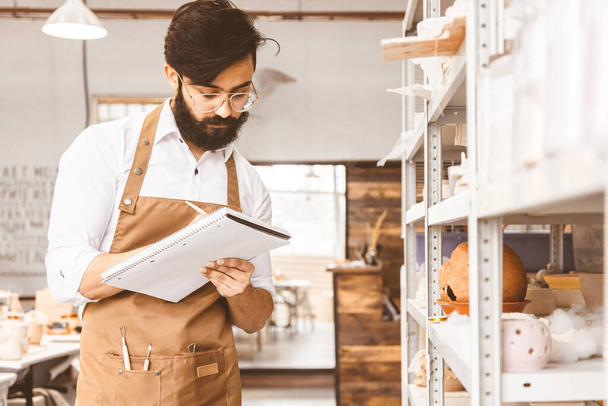 Young attractive businessman a potter with a beard and mustache works in his workshop. Keeps records and transcribes into a notebook inspecting shelves with products, conducts an inventory - Photo, Image