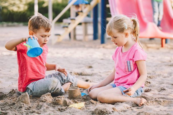 Two Caucasian children sitting in sandbox playing with beach toys. Little girl and boy friends having fun together on playground. Summer outdoor activity for kids. Leisure time lifestyle childhood. - Fotografie, Obrázek