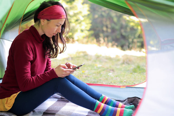 Een Europese vrouw in een tent met uitzicht op het bos houdt een telefoon in haar handen. Een moderne vakantie met apparaten en het internet, om altijd in contact te zijn op vakantie en vakantie. - Foto, afbeelding