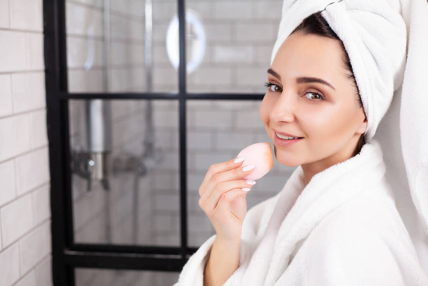 Cute woman in white coat and towel on her head makes herself a makeup - Foto, afbeelding