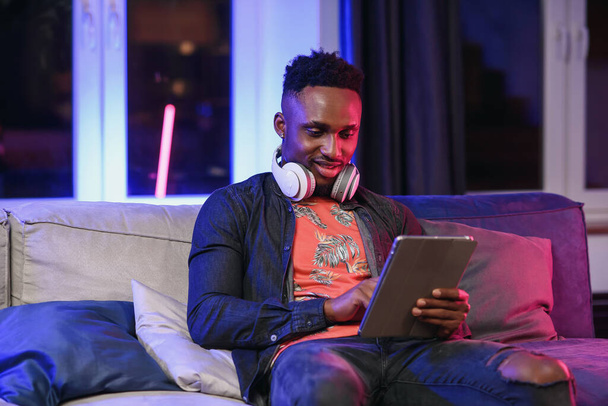 Handsome joyful stylish african american with headphones on his neck relaxing on the soft sofa at home and browsing apps on tablet pc with lucky smile - Foto, immagini