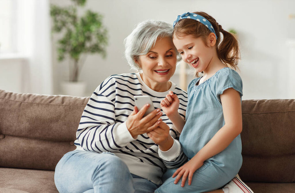 Starší dáma se usmívá a ukazuje média na smartphonu nadšené dívce, zatímco sedí na pohovce a tráví víkend doma - Fotografie, Obrázek