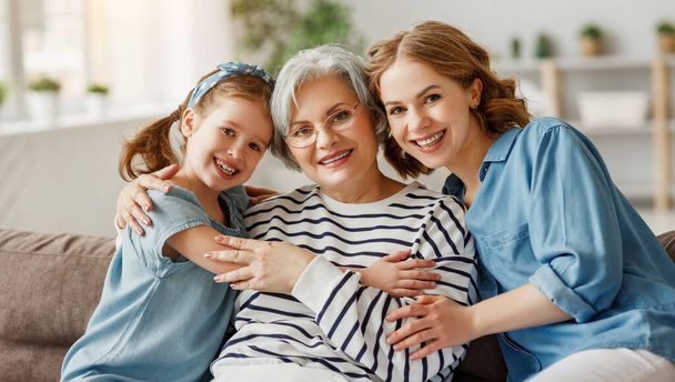Elderly female hugging little girl and young woman smiling and looking at camera while sitting on sofa togethe - Photo, Image