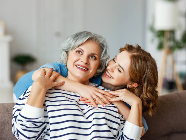 Cheerful young woman hugging aged mother sitting on sofa   while resting together in living room at hom - Фото, изображение