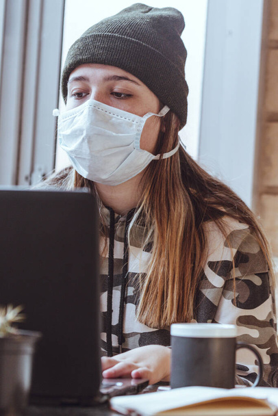 Teen girl with mask protecting herself from the diseases, studying on a terrace with a laptop with a cup of coffee - Fotografie, Obrázek