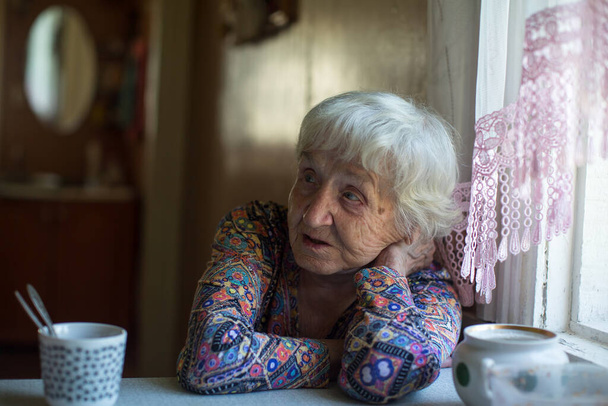 Μια ηλικιωμένη γυναίκα πίνει τσάι καθισμένη στο τραπέζι στο σπίτι της. - Φωτογραφία, εικόνα
