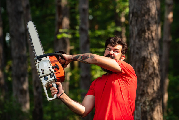 Lumberjack concept. Lumberjack with chainsaw in his hands. Logging. Illegal logging continues today. Lumberjack with chainsaw on forest background. Harvest of timber. - Photo, image
