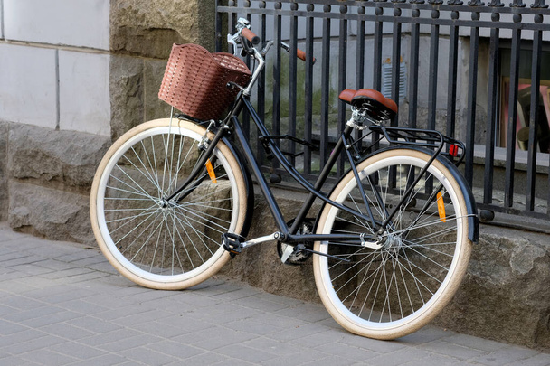 Uma bicicleta de lazer com uma cesta marrom está estacionada perto de uma cerca de metal. Bicicleta da cidade para caminhada e viagem de compras. Roubo de bicicleta fechaduras não confiáveis
. - Foto, Imagem