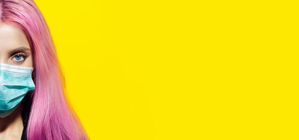 Close-up of half female face, young girl with pink hair and blue eyes, wearing medical flu mask against coronavirus, on yellow background with copy space. - Foto, afbeelding