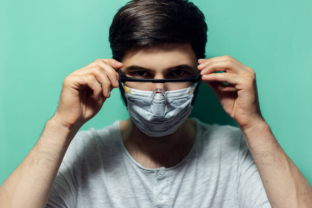 Studio portrait of young man wearing medical flu mask, puts on safety goggles against coronavirus. Isolated on background of aqua menthe color. - Photo, Image