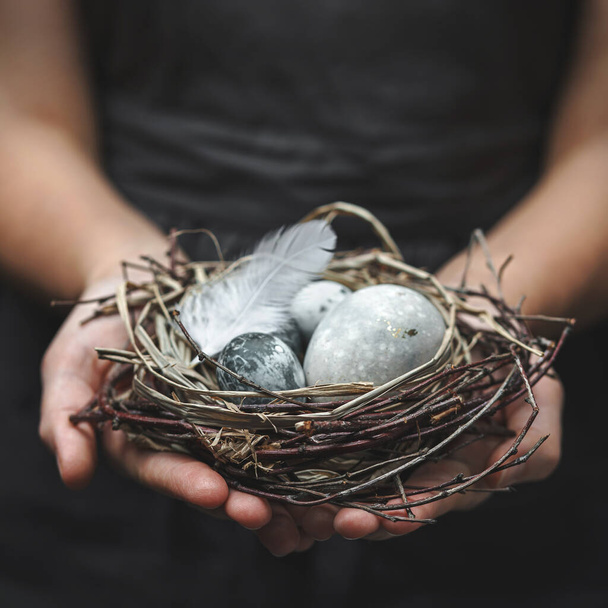Woman hands holding nest with marble Easter eggs with feathers on dark background. Happy Easter holiday, selective focus, toning - Zdjęcie, obraz