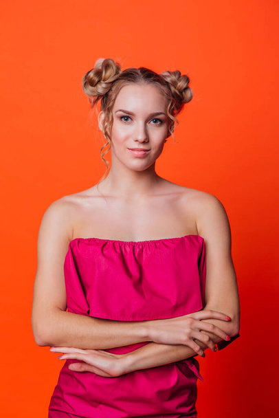 The girl in a red dress on a orange background in the studio. Blonde girl with two hair knots holding hands on chest, looking to the camera and smiling. - Photo, Image