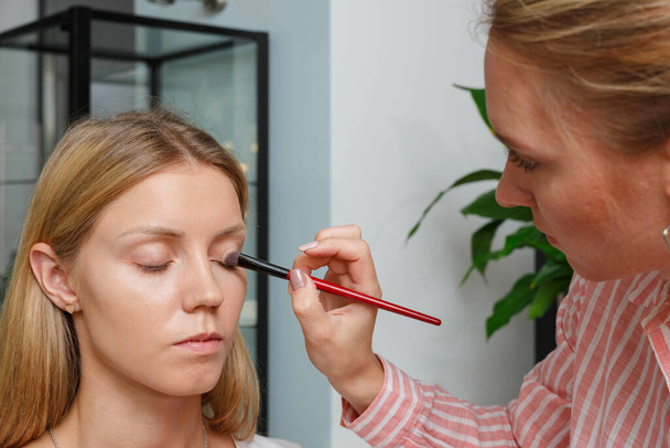 The process of creating makeup. Makeup artist working with a brush on the face of the model. Portrait of a young woman in the interior of a beauty salon. Applying tone to the skin. - Foto, Bild