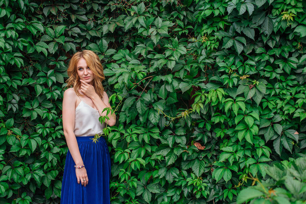 Portrait of a young beautiful woman with make up and curly blond hair, standing next to the wall of wild grape leaves. Copy space. - Fotoğraf, Görsel