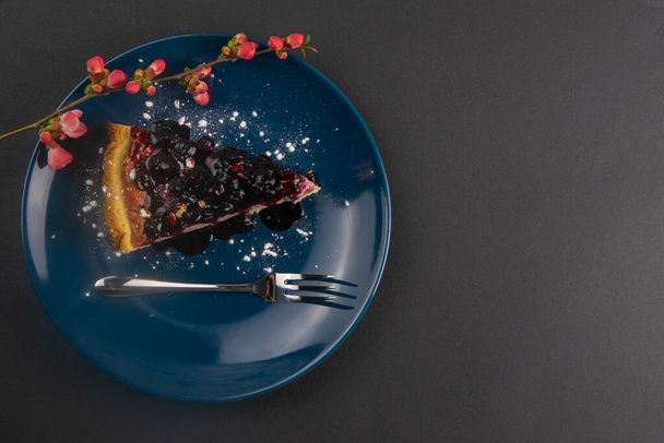 Scheiben von Heidelbeermoussekuchen mit Spiegelglasur dekorierten Beeren und Minzblättern auf einem blauen Teller mit roten Blüten liegen auf schwarzem Tisch, aus nächster Nähe. Top-Ansichten. - Foto, Bild