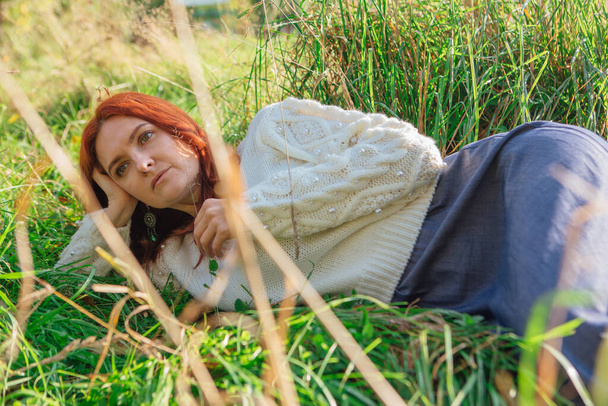 Morning portrait of a beautiful happy woman with red hair laying in grass in white sweater - Photo, image