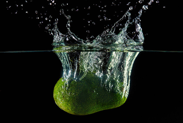 Caída de aguacate en el agua, spray para el diseño, congelación en movimiento. Salpicaduras de agua y verduras aisladas en un fondo negro
. - Foto, imagen