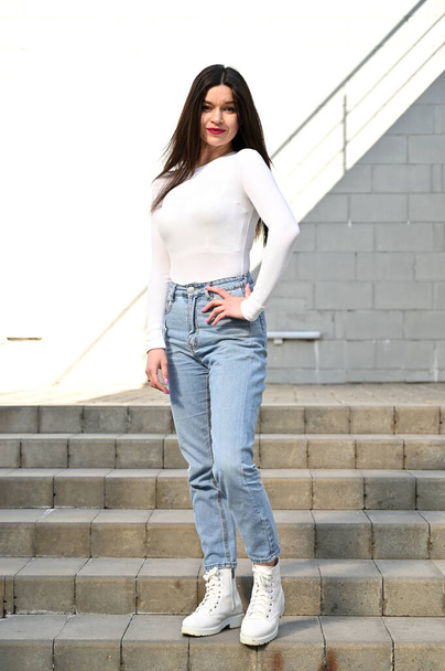 Photo of a caucasian brunette girl in a white blouse and blue jeans stands on the stairs with a smile against the background of a gray wall of a building on a sunny spring day. - Фото, изображение