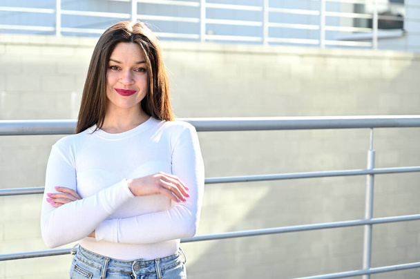 Caucasian portrait of a pretty brunette girl in a white blouse and blue jeans stands near the railing with a smile against the background of a gray wall of a building on a sunny spring day. - Фото, изображение