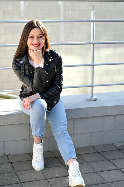 Vertical Caucasian portrait of a lovely brunette girl in a black jacket and blue jeans with a smile sits on the railing on a sunny spring day. - Foto, immagini