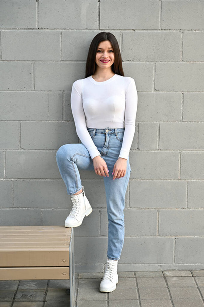 Vertical Caucasian portrait of a pretty brunette girl in a white blouse and blue jeans stands near the wall of a building on a spring day in the city. Model is happy to pose. - Фото, зображення