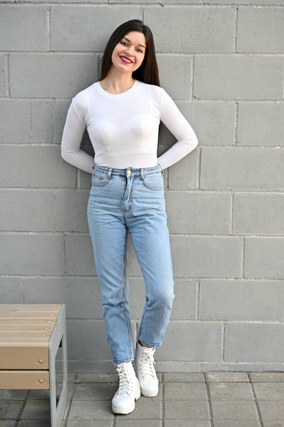 Vertical Caucasian portrait of a pretty brunette girl in a white blouse and blue jeans stands near the wall of a building on a spring day in the city. Model is happy to pose. - Valokuva, kuva