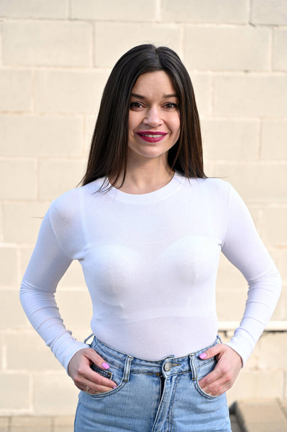 Vertical Caucasian portrait of a pretty brunette girl in a white blouse and blue jeans stands near the wall of a building on a spring day in the city. Model is happy to pose. - Фото, изображение