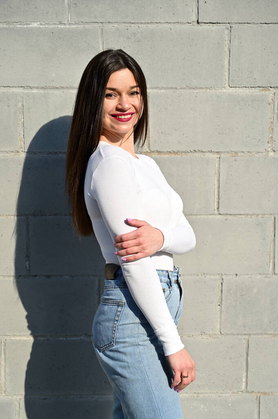 Vertical Caucasian portrait of a pretty brunette girl in a white blouse and blue jeans stands near the wall of a building on a spring day in the city. Model is happy to pose. - Фото, изображение