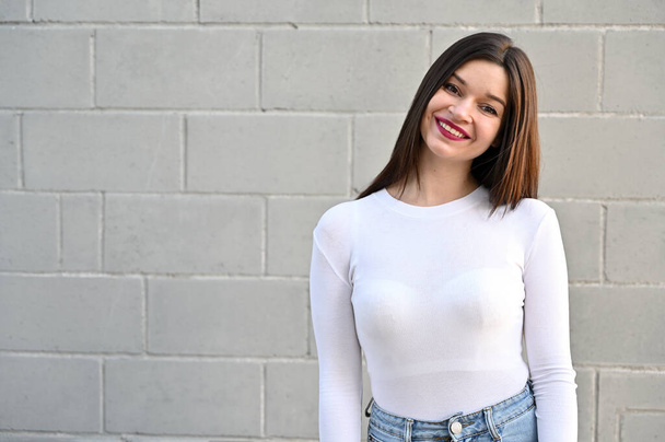 A caucasian portrait of a pretty brunette girl in a white blouse stands near the wall of a building on a spring day in the city. Model with pleasure and smile posing. - Photo, image