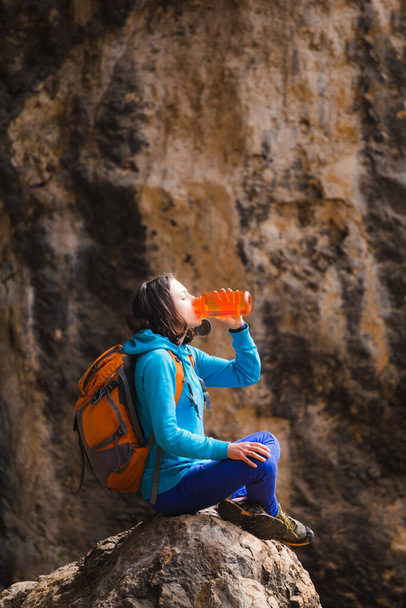 Žena s batohem pije vodu z opakovaně použitelné láhve. Dívka sedí na velkém kameni a uhasí žízeň. Cestovatel na pozadí skal. - Fotografie, Obrázek
