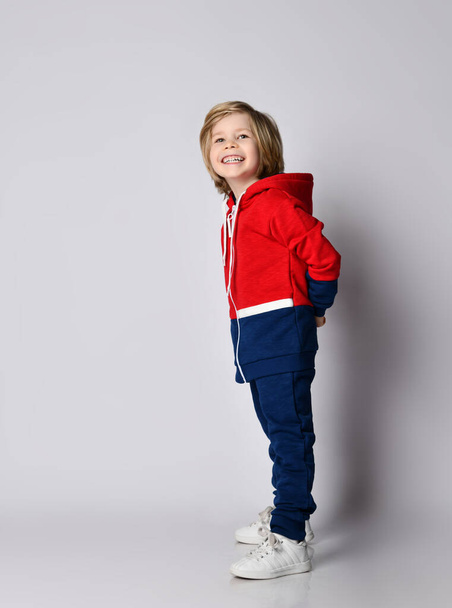 Cheerful laughing frolic blond kid boy in blue and red hoodie and pants holds hands behind back, hiding something - Foto, Imagem