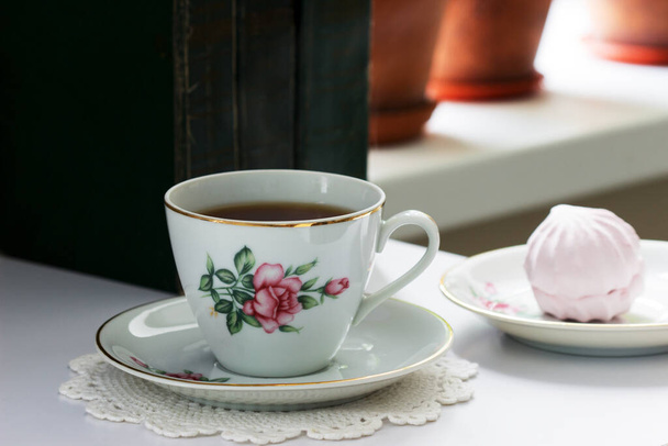 ヴィンテージのカップの中のお茶、ヴィンテージのソーサーの上のマシュマロ、そして軽い背景の古い本. - 写真・画像