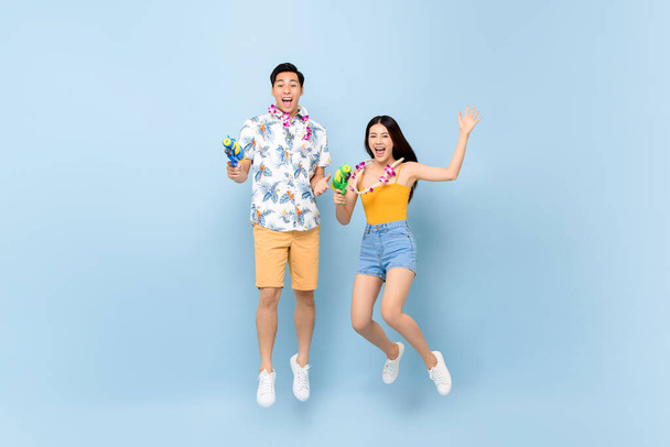 Young Asian couple in summer outfits with water guns jumping in studio blue background for Songkran festival in Thailand and southeast Asia - Photo, Image
