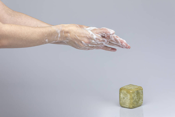 Las manos de los blancos se lavan unas a otras con jabón verde espumoso de Marsella aislado frente a un fondo gris con luces y sombras estructurantes
 - Foto, imagen