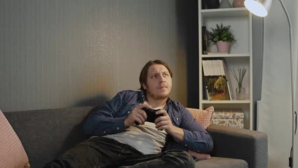 Young man lays on a couch plays games, holds a joystick in his hands - Imágenes, Vídeo