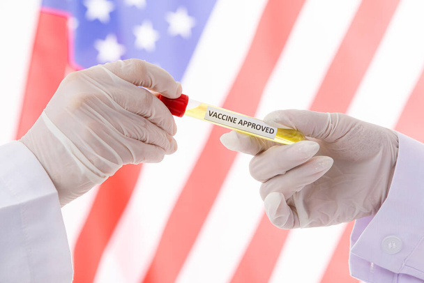 Zwei Hände von Ärzten oder Forschern liefern Reagenzgläser mit der Aufschrift Impfstoffzulassung, im Hintergrund ist die Flagge der Vereinigten Staaten verschwommen. Konzept für Gesundheitsversorgung und Bekämpfung des Virus-Ausbruchs. - Foto, Bild